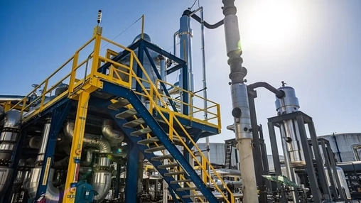 Industrial refinery with a blue steel platform, yellow safety railings, distillation towers, and pipework.