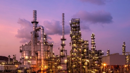 Refinery towers and industrial pipework lit by floodlights at dusk against a vivid purple and pink sunset sky